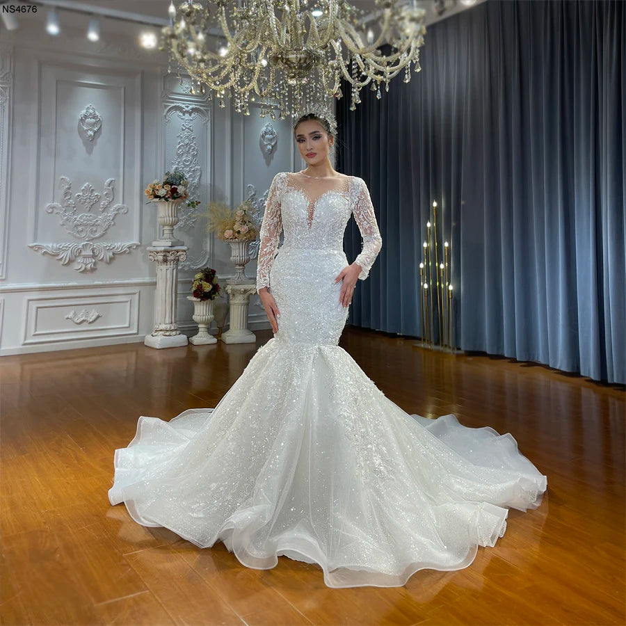 Full-length front view of a model wearing an elegant lace mermaid wedding dress with long sleeves, a beaded illusion neckline, and a voluminous romantic train in a luxury bridal showroom.