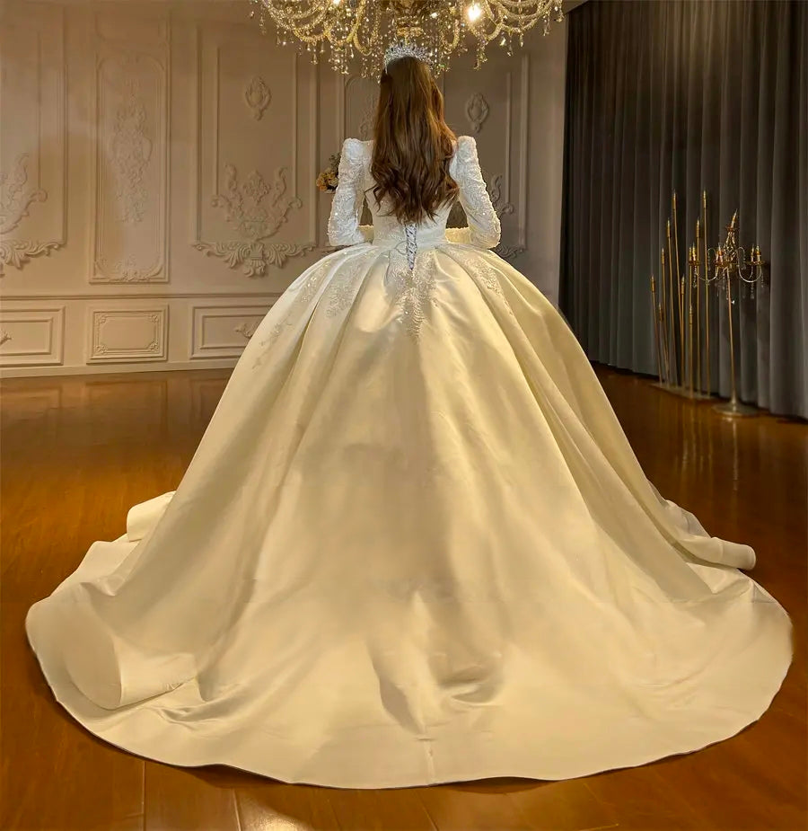 Back view of a stunning modest bridal ball gown emphasizing the grand, flowing silhouette and premium satin fabric under a chandelier.
