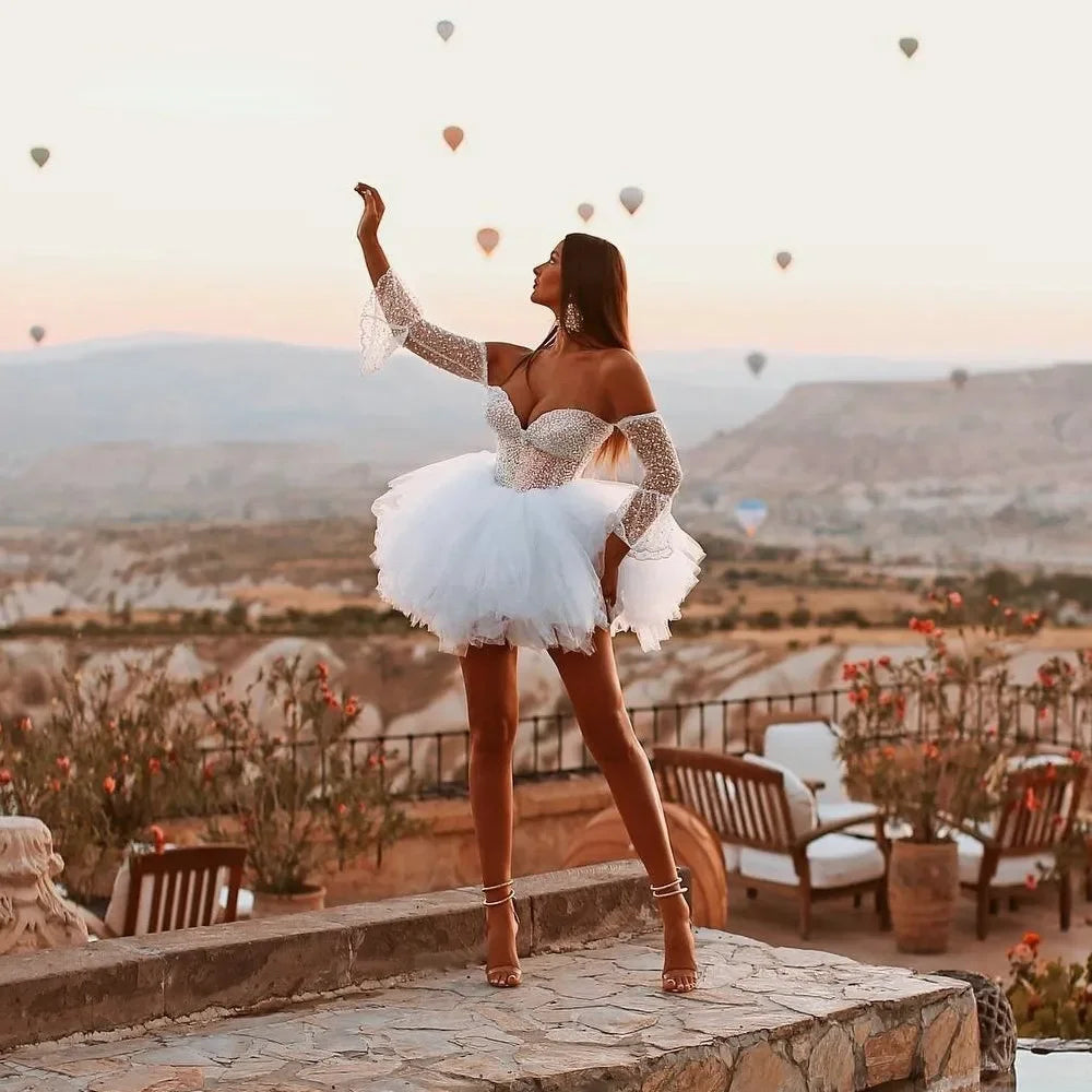 Modern beach wedding reception dress with an off-shoulder neckline, embellished long sleeves, and a short, fluffy A-line organza skirt.