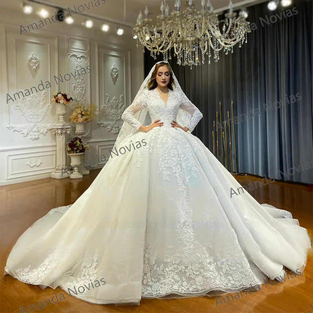 Model wearing an elegant off-white bridal ball gown featuring a plunging V-neckline, long sheer lace sleeves, and a voluminous sparkling skirt under a crystal chandelier.