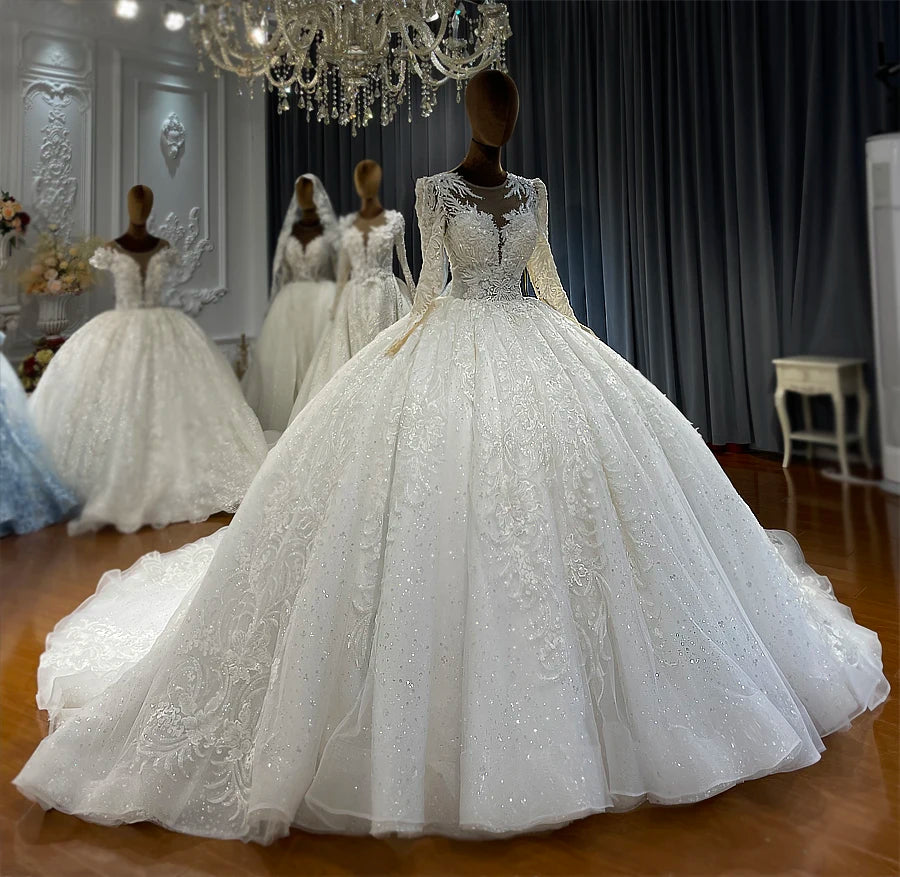 Mannequin display of a stylish off-white bridal gown with long illusion sleeves and a voluminous glitter tulle skirt adorned with silver embroidery.