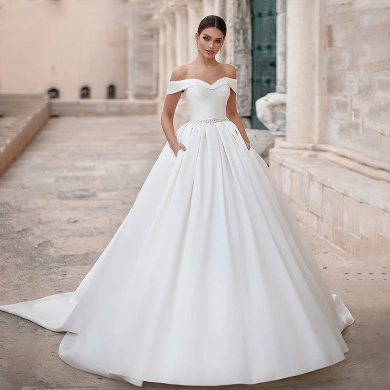 Elegant satin princess wedding gown featuring a sweetheart neckline, off-the-shoulder cap sleeves, and a sparkling crystal sash.
