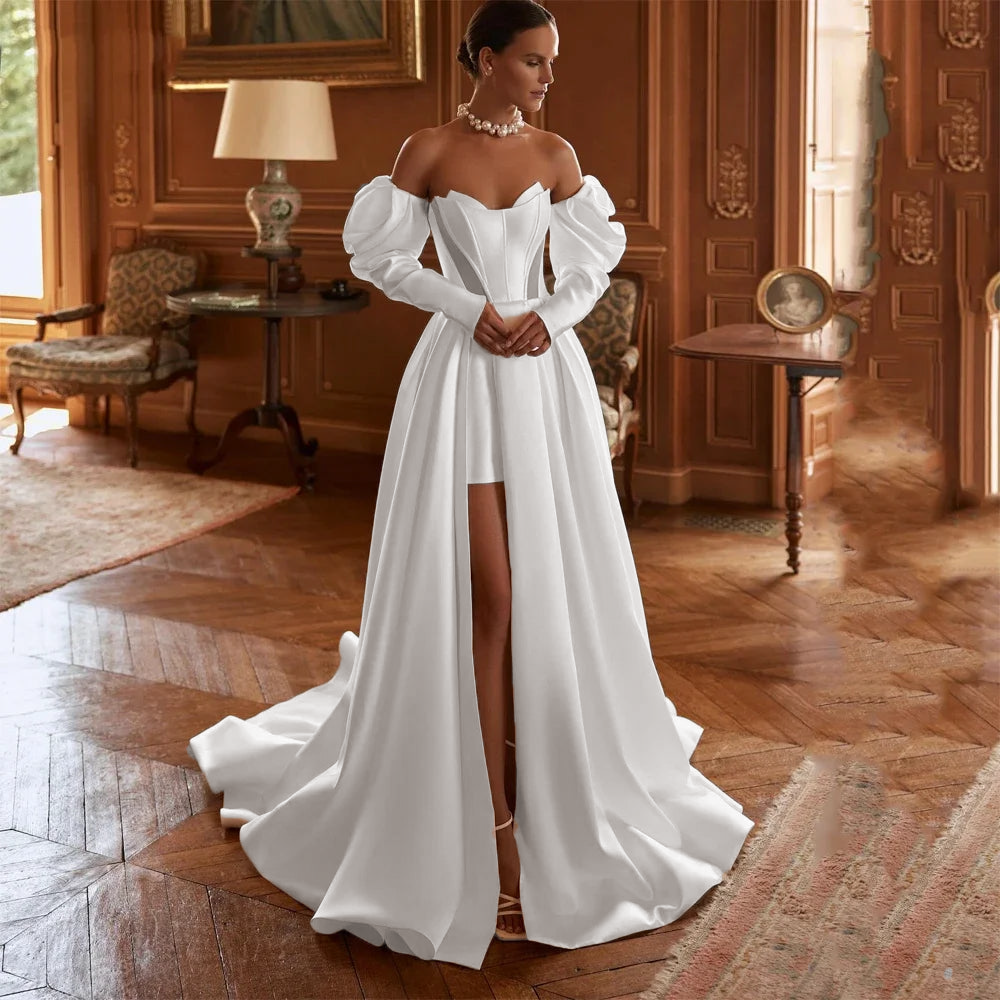 A front-facing portrait of a bride modeling a luxury satin A-line bridal gown with off-the-shoulder sleeves, a high slit, and a voluminous skirt.