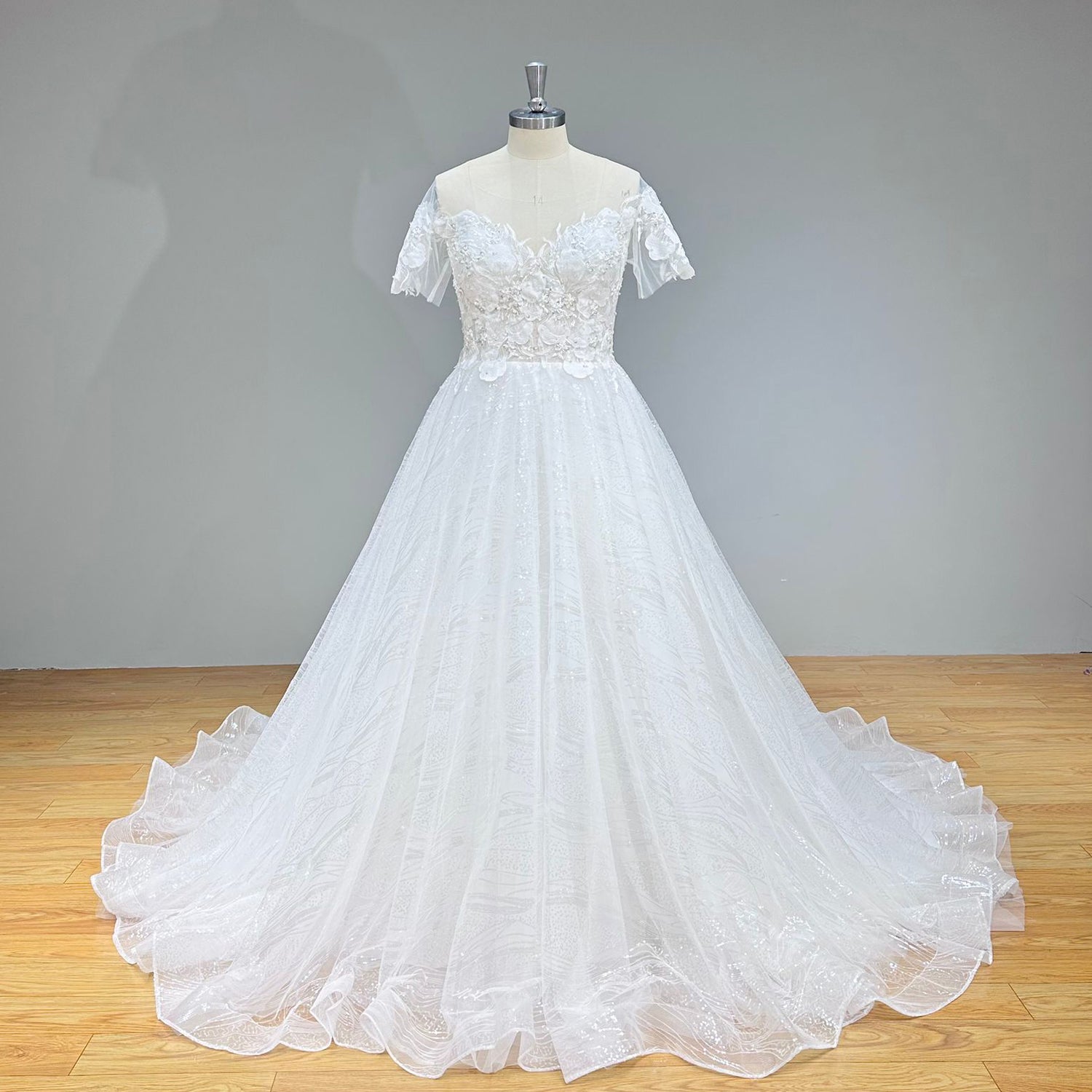 Studio front display of an off-the-shoulder short sleeve wedding gown featuring a shimmering sequin tulle skirt and floral applique details.