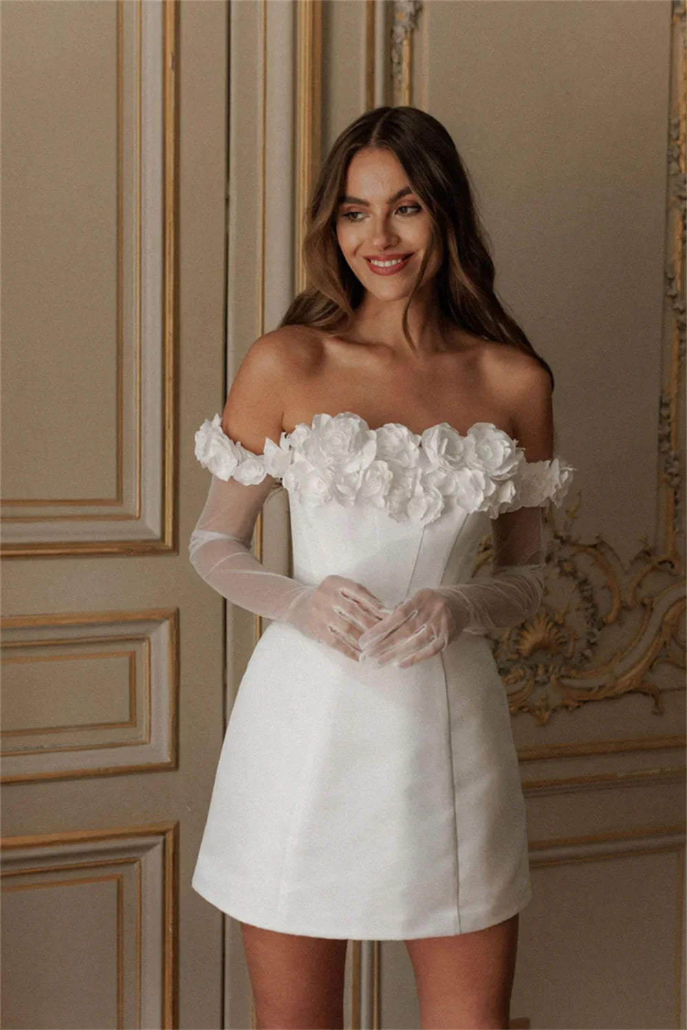 Model posing in a white off-the-shoulder mini wedding dress with intricate 3D floral details and a high side leg slit.
