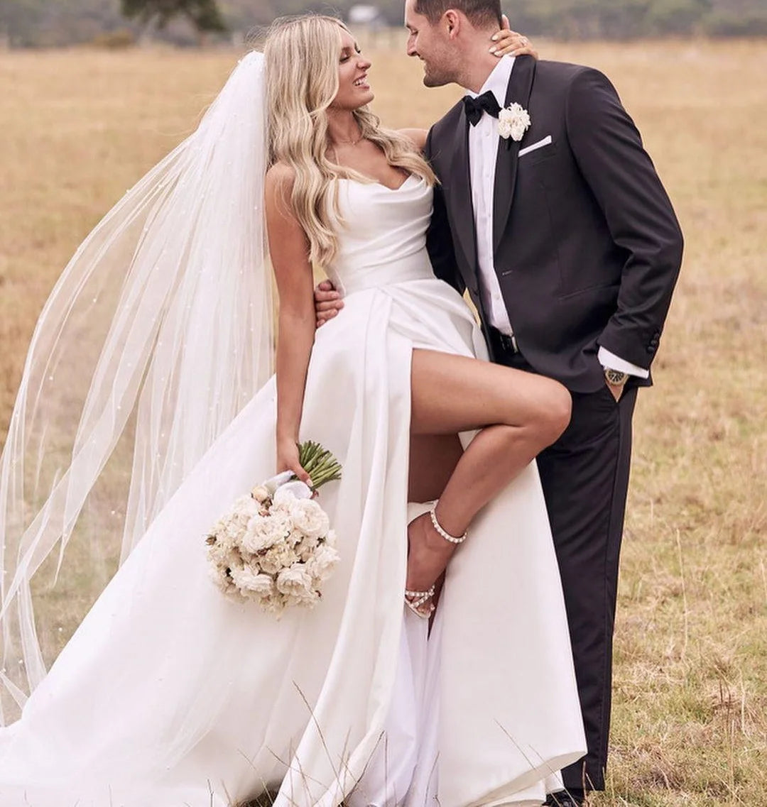 Bride in a simple satin strapless wedding dress with a high side slit, embracing her groom in a field.