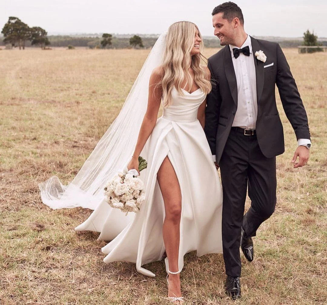 A couple walking through a field, the bride wearing an elegant sleeveless satin wedding gown featuring a thigh-high slit and a sweep train.