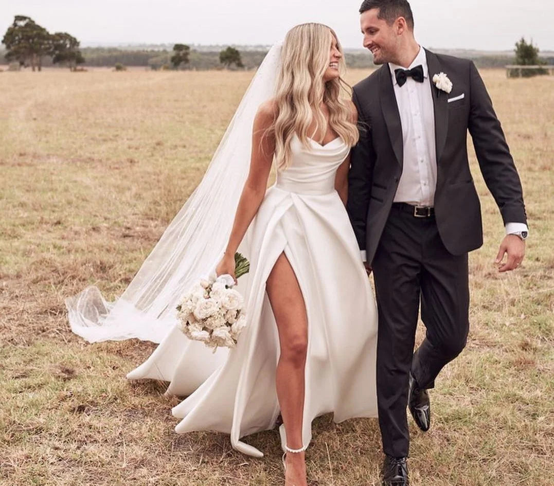 Beautiful bride in a strapless satin vintage-style wedding dress walking outdoors with her groom, highlighting the high slit and sweep train.