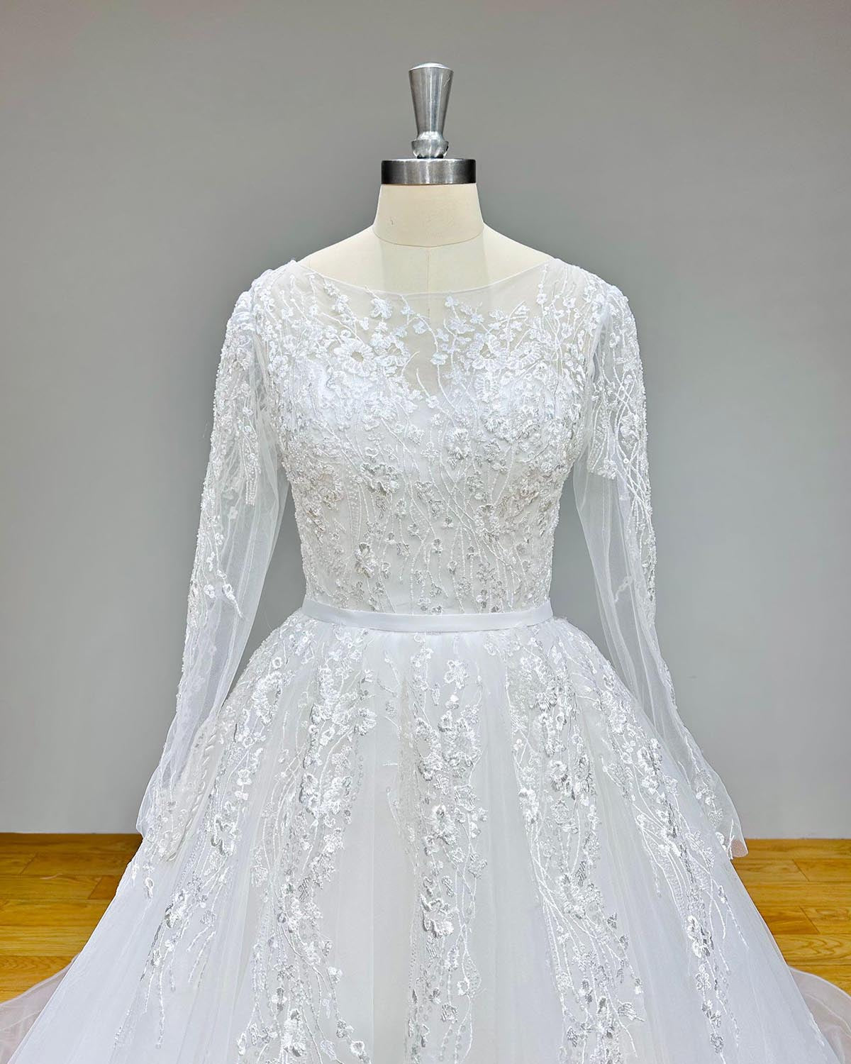 Close-up view of the bodice of an A-line tulle wedding dress featuring long sleeves, intricate lace embroidery, and a satin waist belt.