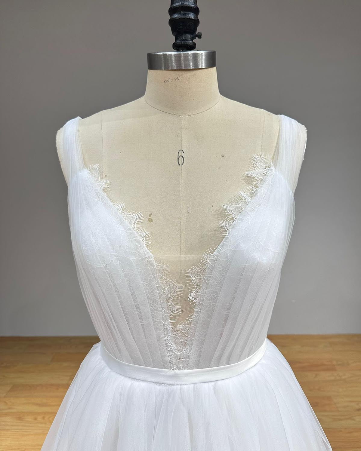 Macro close-up of a pleated tulle bridal bodice featuring a sheer mesh illusion insert and a fine eyelash lace border along the plunging neckline on a mannequin.