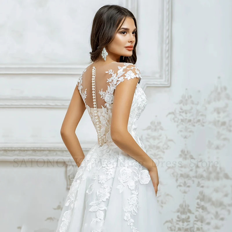Rear profile view of a short lace bridal gown showcasing a sheer illusion back adorned with floral embroidery and a classic vertical row of pearl button closures.