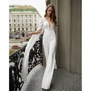 Full-length front view of a model on a decorative balcony wearing an elegant white wedding jumpsuit with a plunging deep V-neckline, structured shoulders, and dramatic floor-length split cape sleeves.