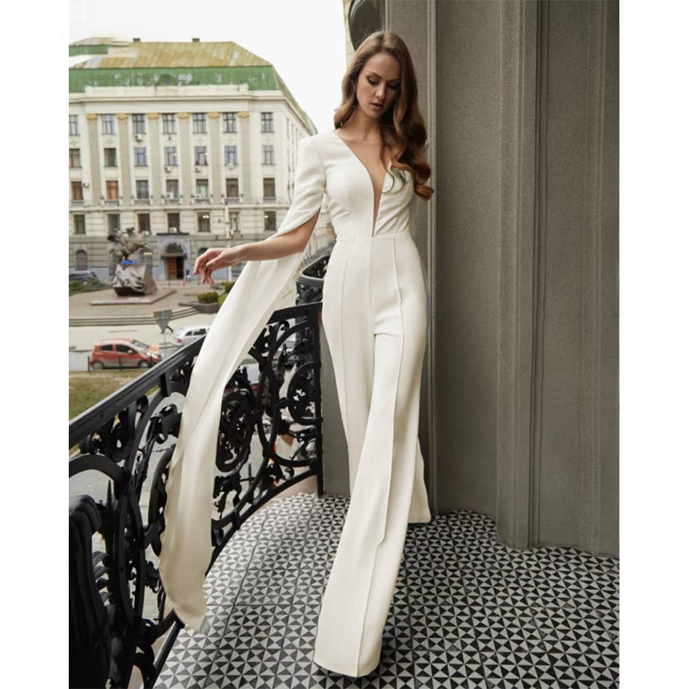 Luxury bridal gown pants set featuring a modern plunging V-neck white jumpsuit bodice and fluid wide-leg trousers paired with sweeping, long cape sleeves.