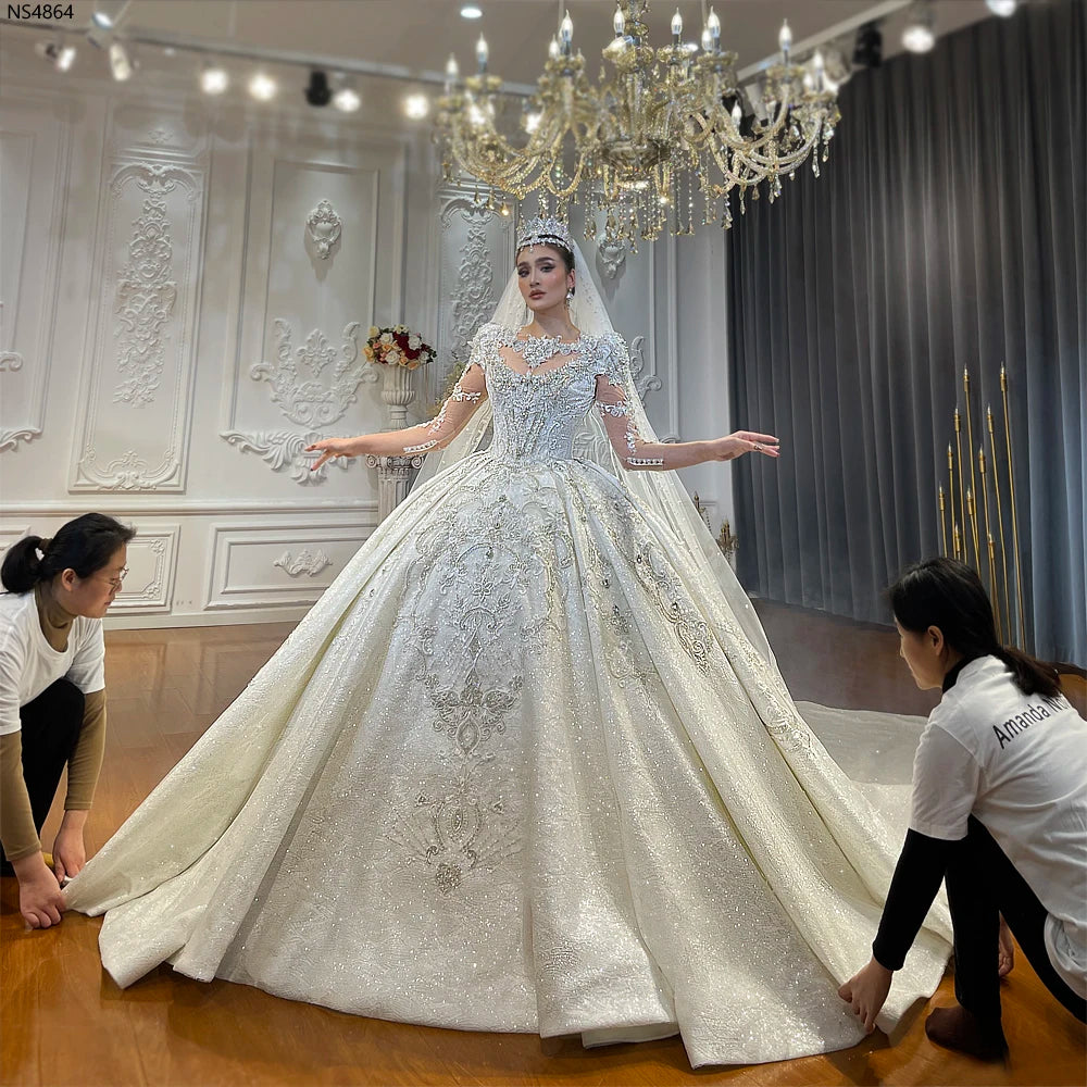 Front view of a bride wearing a luxury empire wedding dress with a high neck and long sleeves, featuring heavy crystal beading and a shimmering grand ball gown skirt.