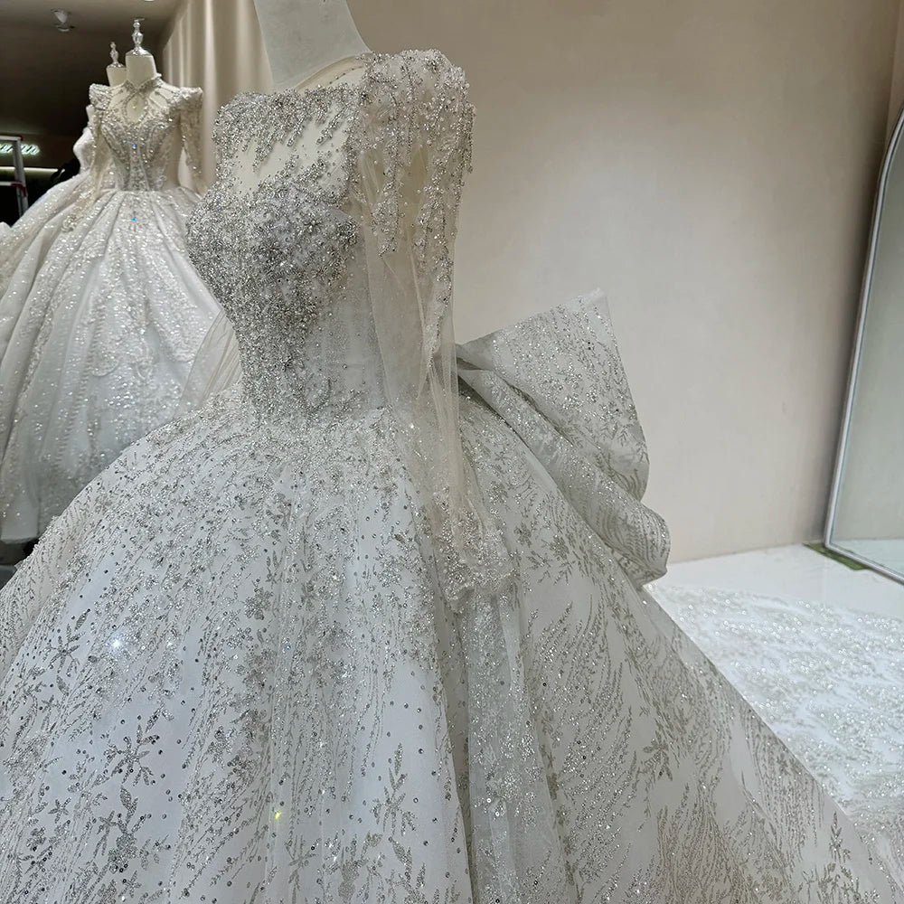 Side close-up of a luxury wedding gown bodice on a mannequin, highlighting the sheer long sleeves with beaded cuffs, structured shoulders, and the profile of a massive back bow.