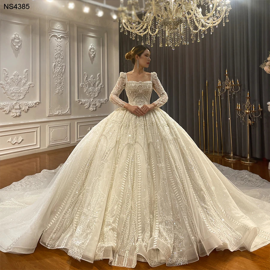 Full front view of a fantasy square collar ball gown wedding dress featuring long sleeves, heavy crystal beading, and a voluminous shimmering skirt in a luxury bridal studio.