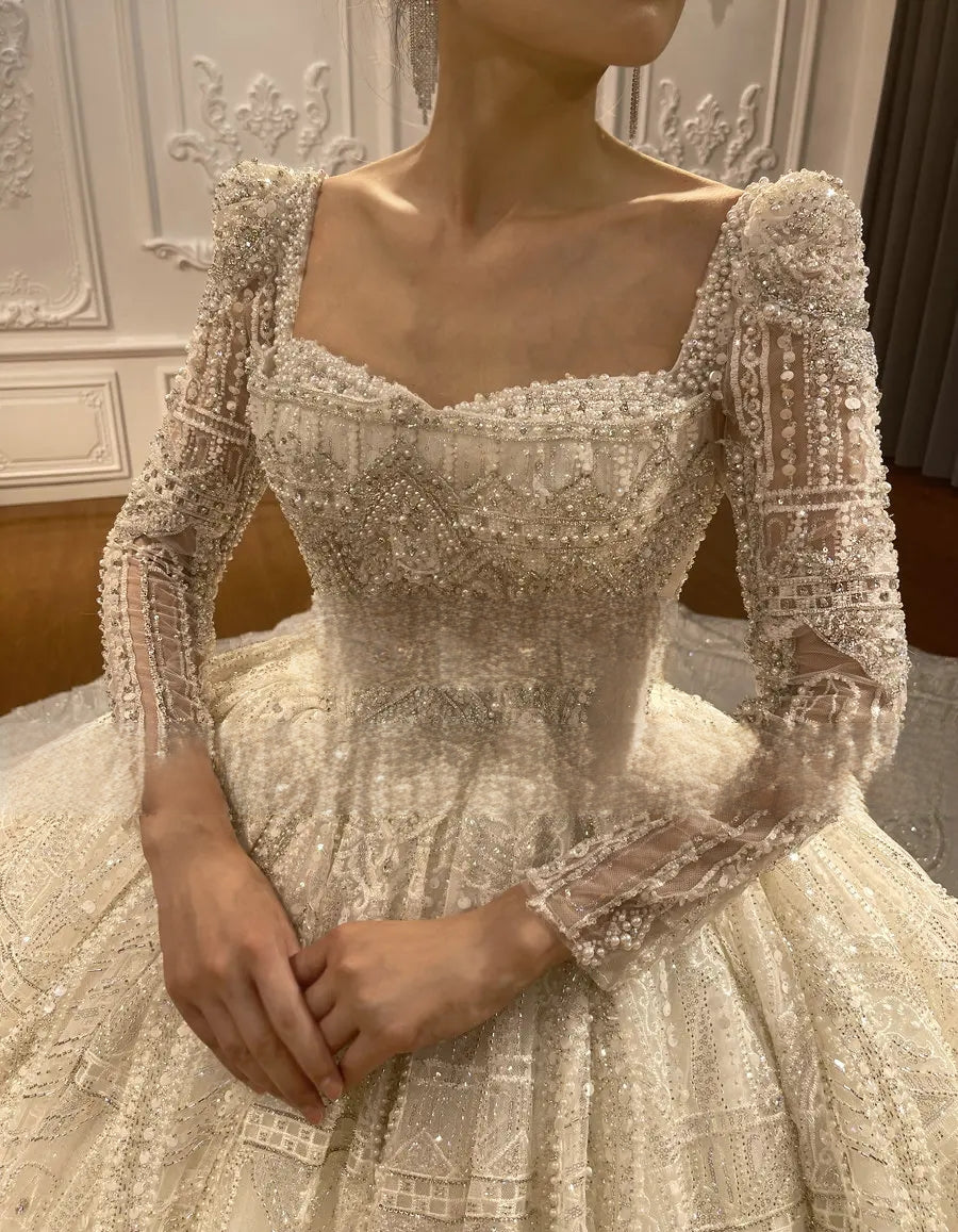 Close-up of a luxury wedding dress bodice showing the intricate hand-beaded square neckline and delicate semi-sheer long sleeves with sparkle detailing.