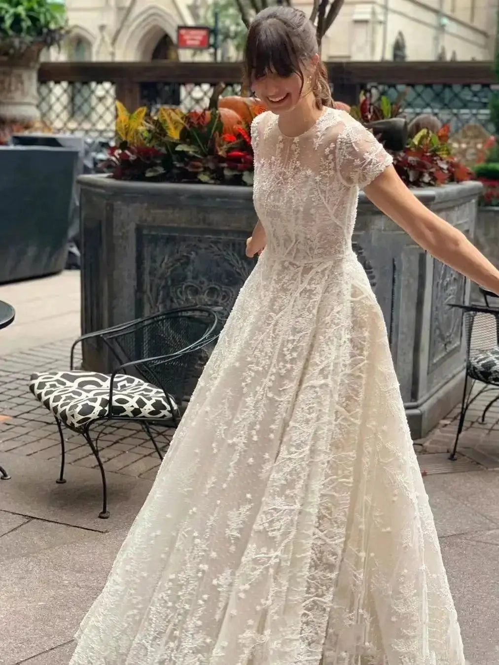 A joyful bride wearing a floor-length boho-inspired wedding dress with lace appliqués and short sleeves, standing outdoors.