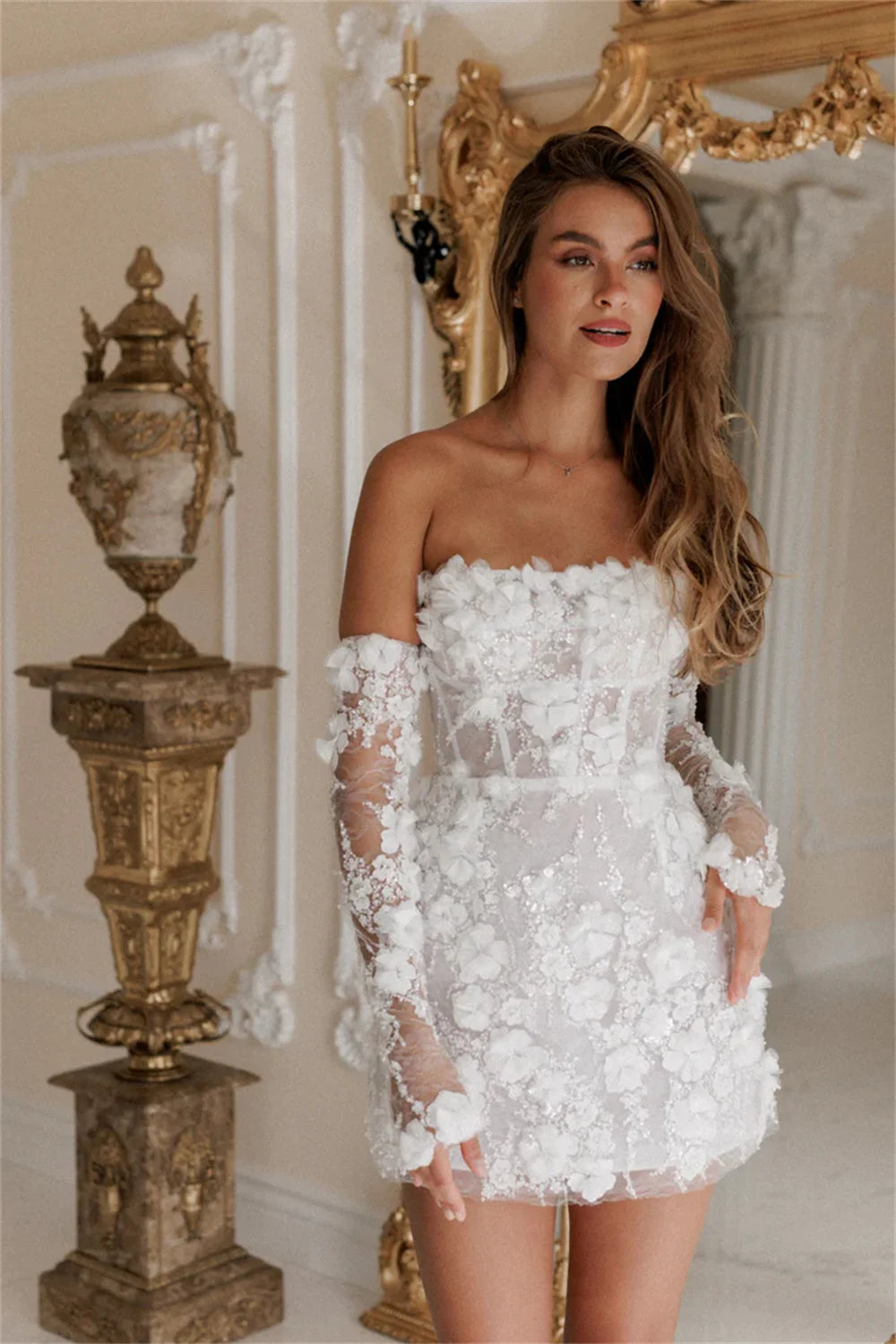 A model standing in a white 3D floral applique mini dress with detachable lace sleeves, displayed in an elegant indoor setting with a golden mirror.