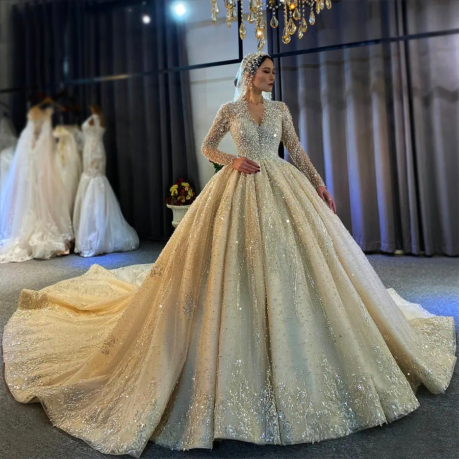Front angled view of an elegant model wearing a luxury ball gown wedding dress featuring long sleeves and a deep V-neckline, heavily embellished with full pearls and crystals.