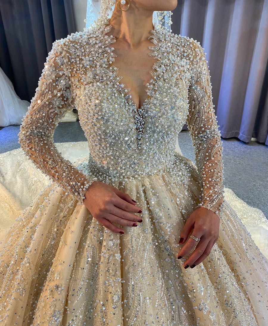Close-up detail of the front bodice of a luxury wedding gown showcasing handcrafted pearl work, shimmering crystals, and elegant long sleeves on a V-neck silhouette.