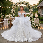 Smiling bride wearing a luxury white ball gown wedding dress with a plunging sweetheart neckline, dramatic side ruffles, and off-the-shoulder straps at a beautiful outdoor garden venue.