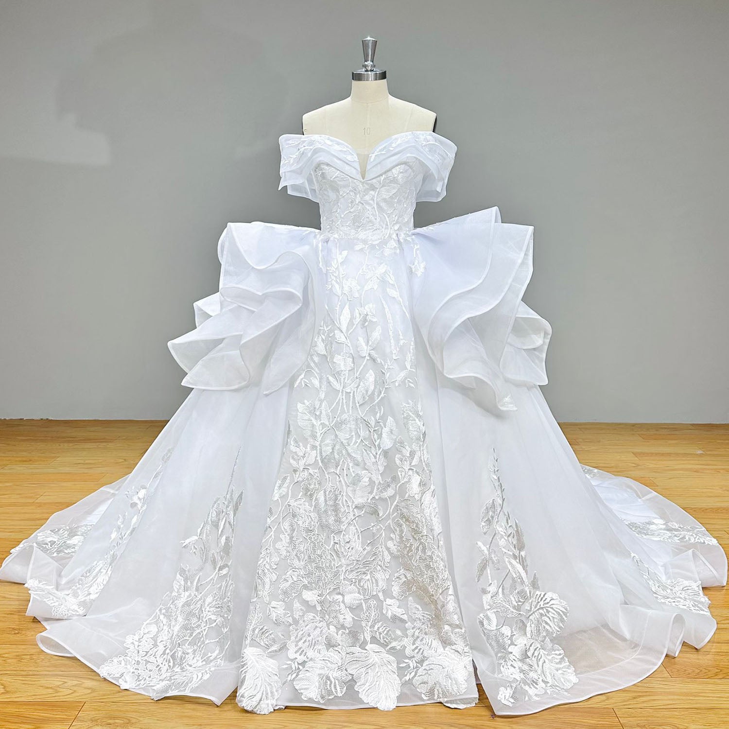 Full front product photography of the architectural off-the-shoulder ball gown wedding dress with dramatic side ruffles on a mannequin against a gray studio background.
