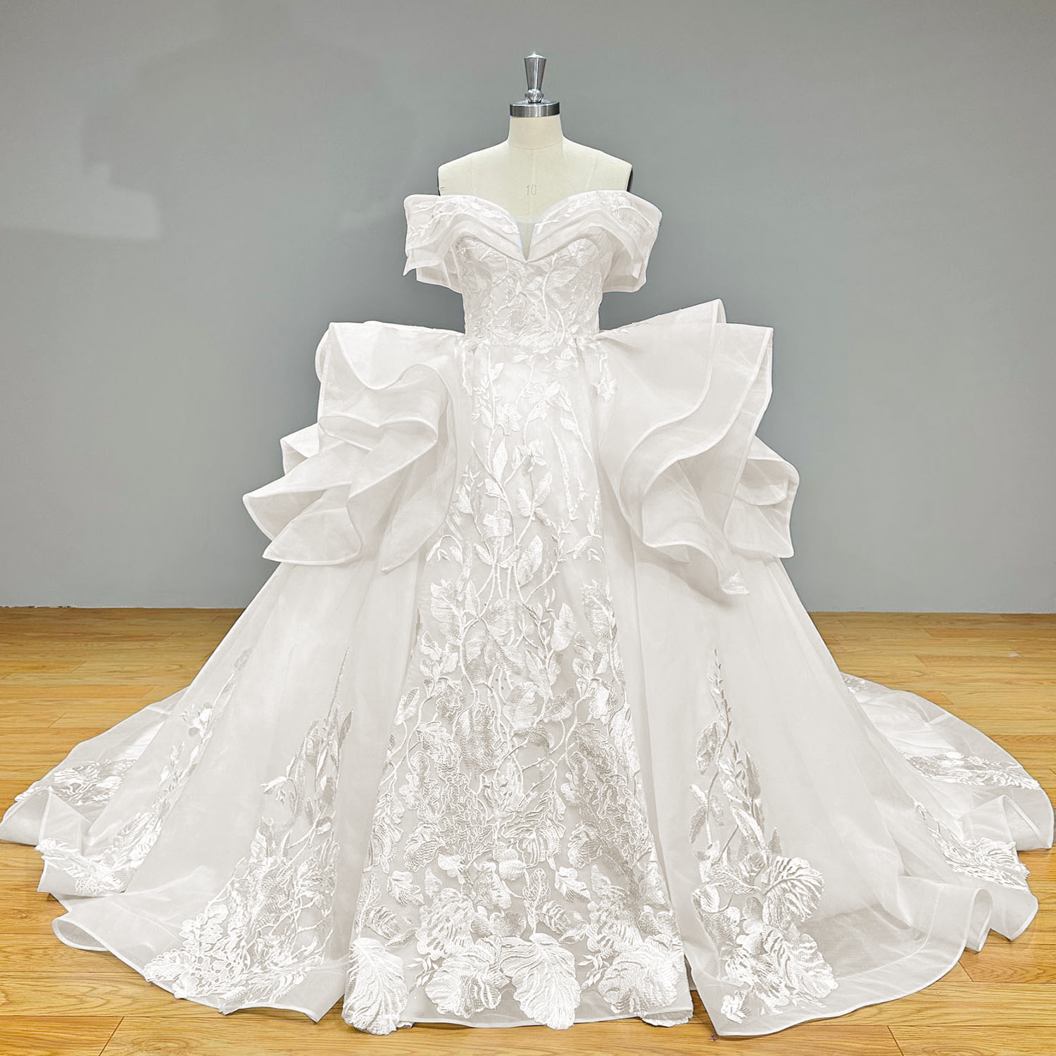 Full front product photography of the architectural off-the-shoulder ball gown wedding dress with dramatic side ruffles on a mannequin against a gray studio background.