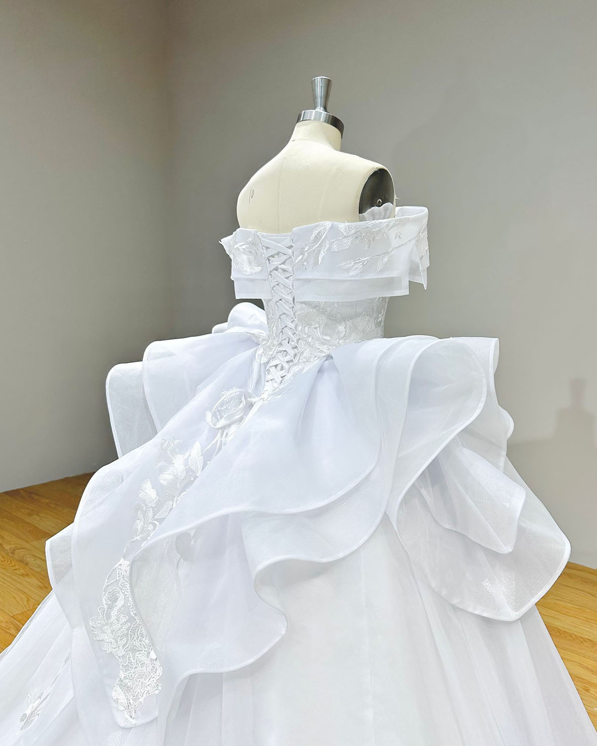 Three-quarter back angle of the wedding dress bodice on a mannequin, showing the adjustable satin ribbon corset back and voluminous layered ruffles on the skirt hips.
