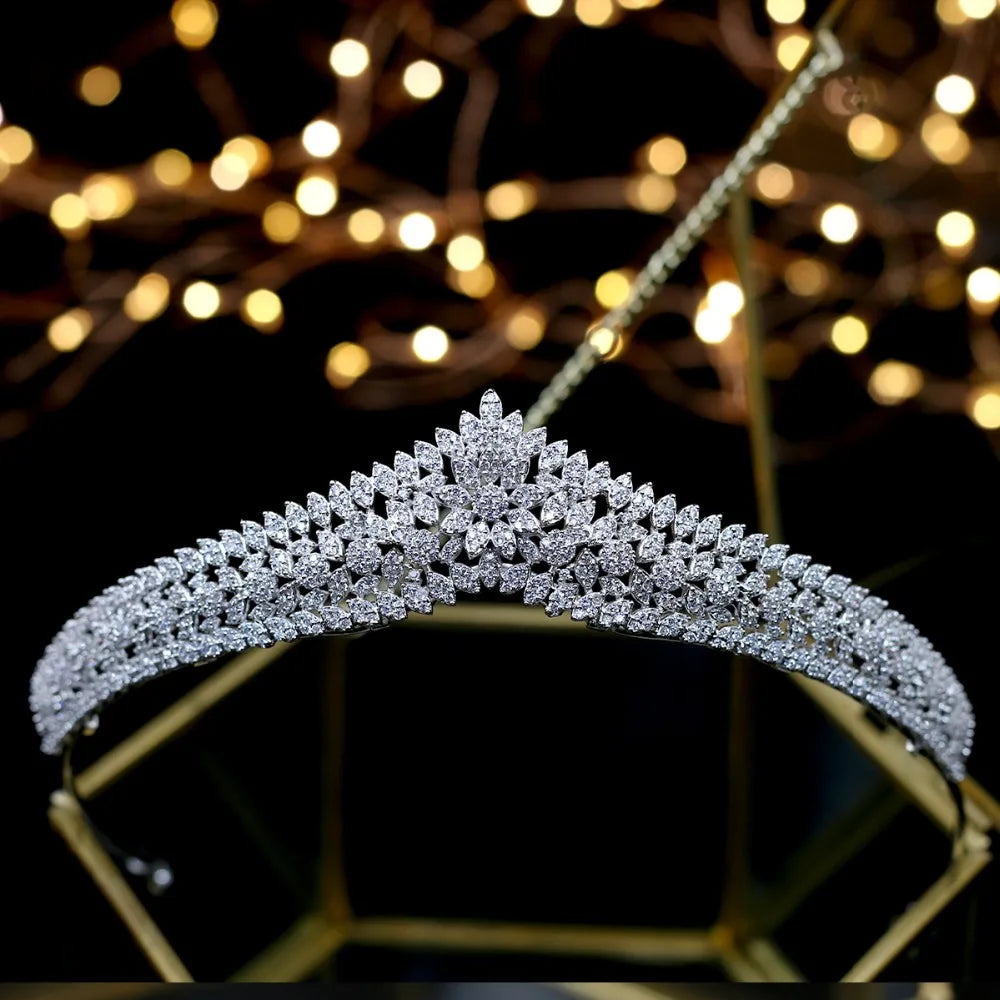 Front profile of a luxury nupcial bridal tiara sparkling with round and marquise-cut crystals, displayed on a gold geometric stand with background bokeh.