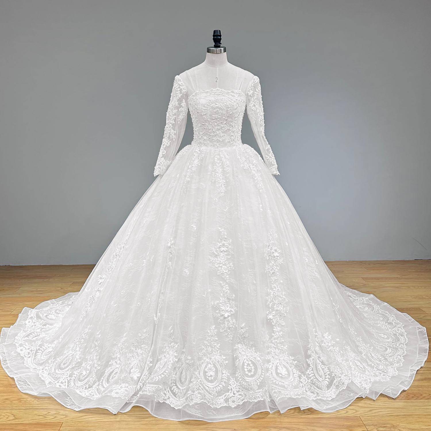 Studio front perspective of the heavy baroque style embroidered white lace wedding gown with long sleeves on a mannequin against a solid gray background.