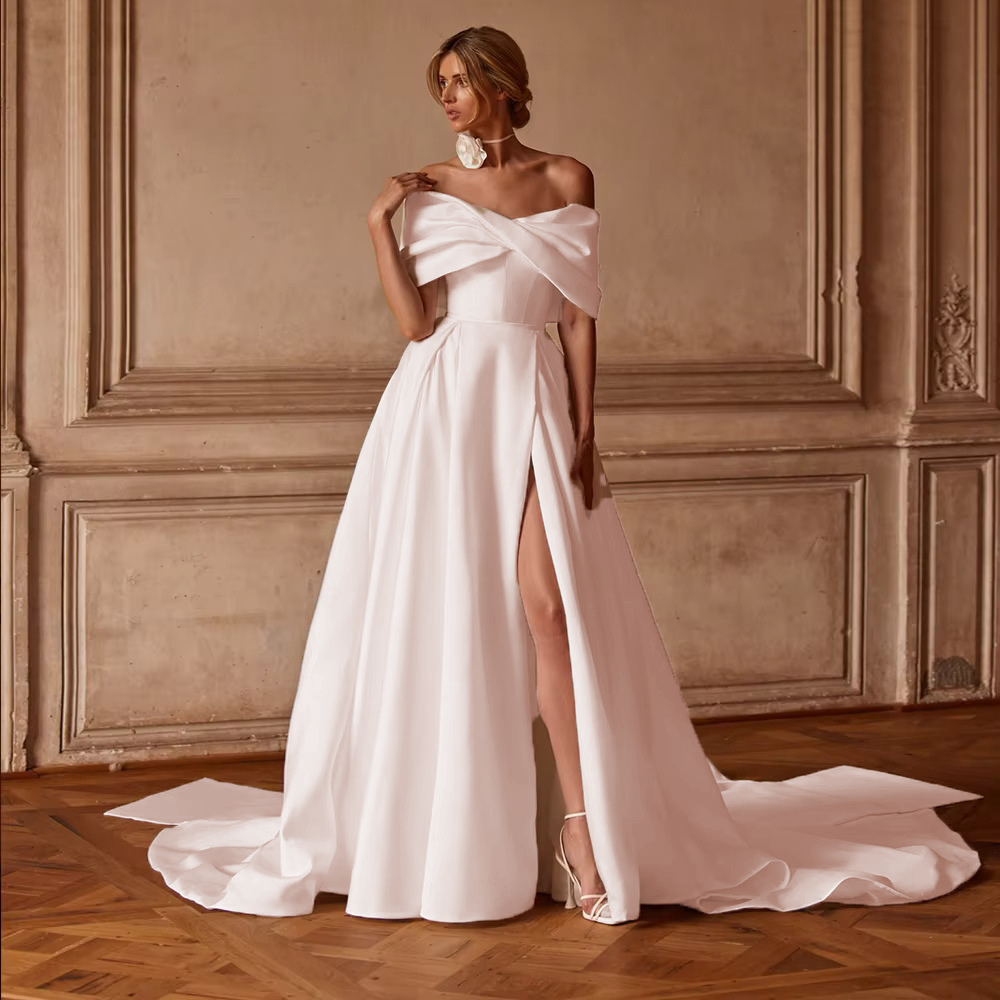Romantic soft blush pink satin A-line wedding dress featuring an architectural draped off-the-shoulder portrait collar and a flowing slit skirt.