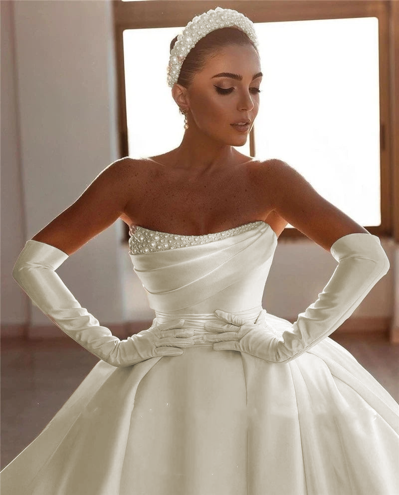 An elegant studio shot of a bride showcasing a strapless princess wedding dress with a pearl-embellished neckline and long satin gloves.