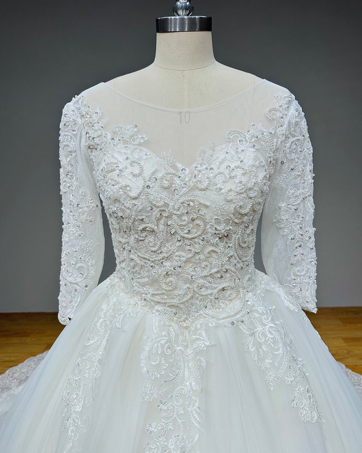 Close-up front view of a bridal bodice on a mannequin, highlighting the sheer illusion scoop neckline, heavy crystal beadwork, pearl appliques, and intricate scroll lace embroidery.