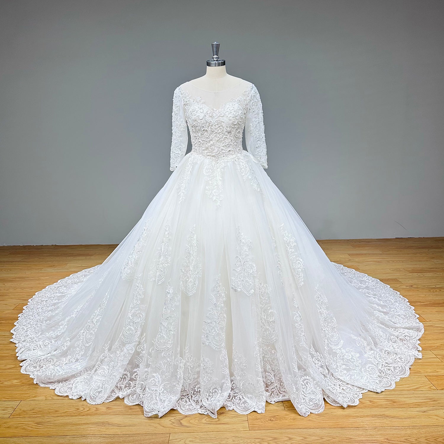 Front product display of a classic tulle ball gown bridal dress with embroidered lace appliques and sheer half sleeves against a gray wall backdrop.