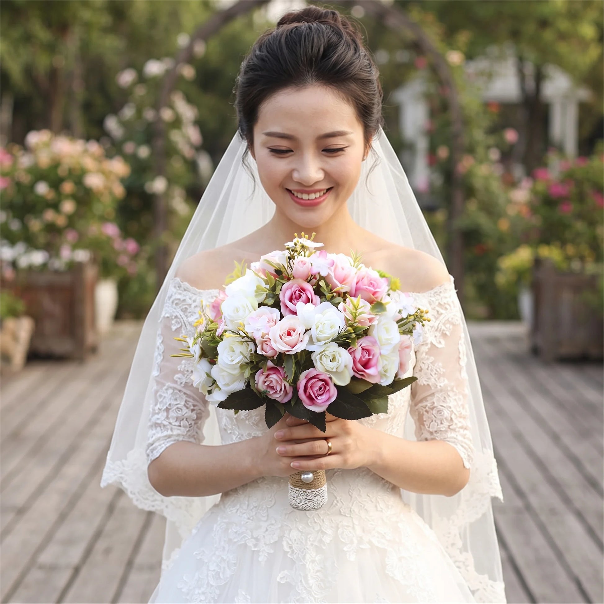 Medium portrait of a smiling bride wearing an off-the-shoulder lace dress and veil, holding a handmade wedding bridal bouquet composed of realistic pink, white, and cream silk roses with a lace-wrapped stem.