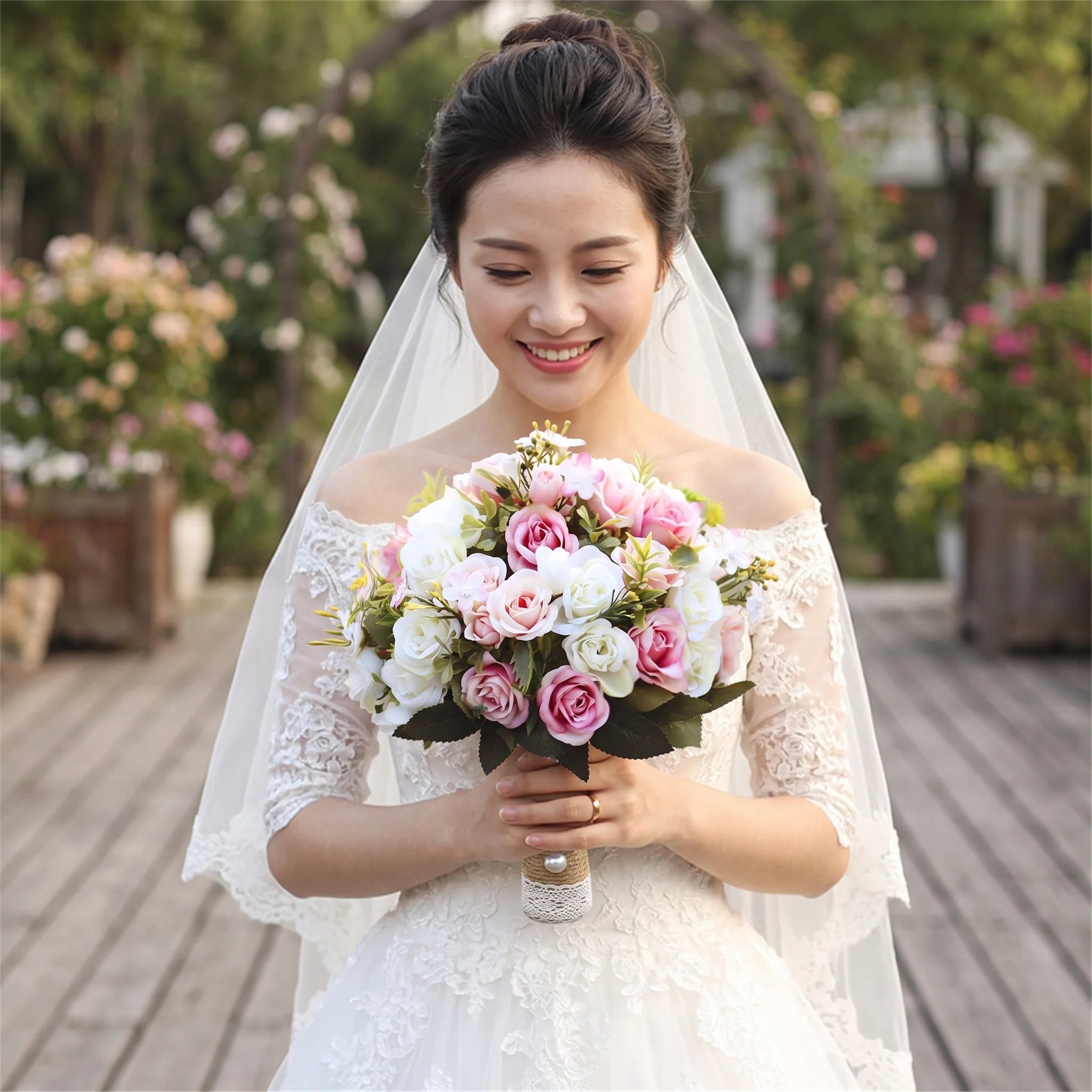 Medium portrait of a smiling bride wearing an off-the-shoulder lace dress and veil, holding a handmade wedding bridal bouquet composed of realistic pink, white, and cream silk roses with a lace-wrapped stem.
