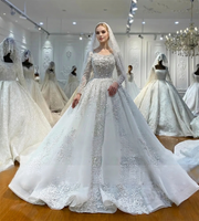 Full-length front view of an elegant model in a heavy beaded pearl wedding dress with long sleeves and a matching beaded veil, showcasing a luxury off-white ball gown silhouette in a bridal boutique.