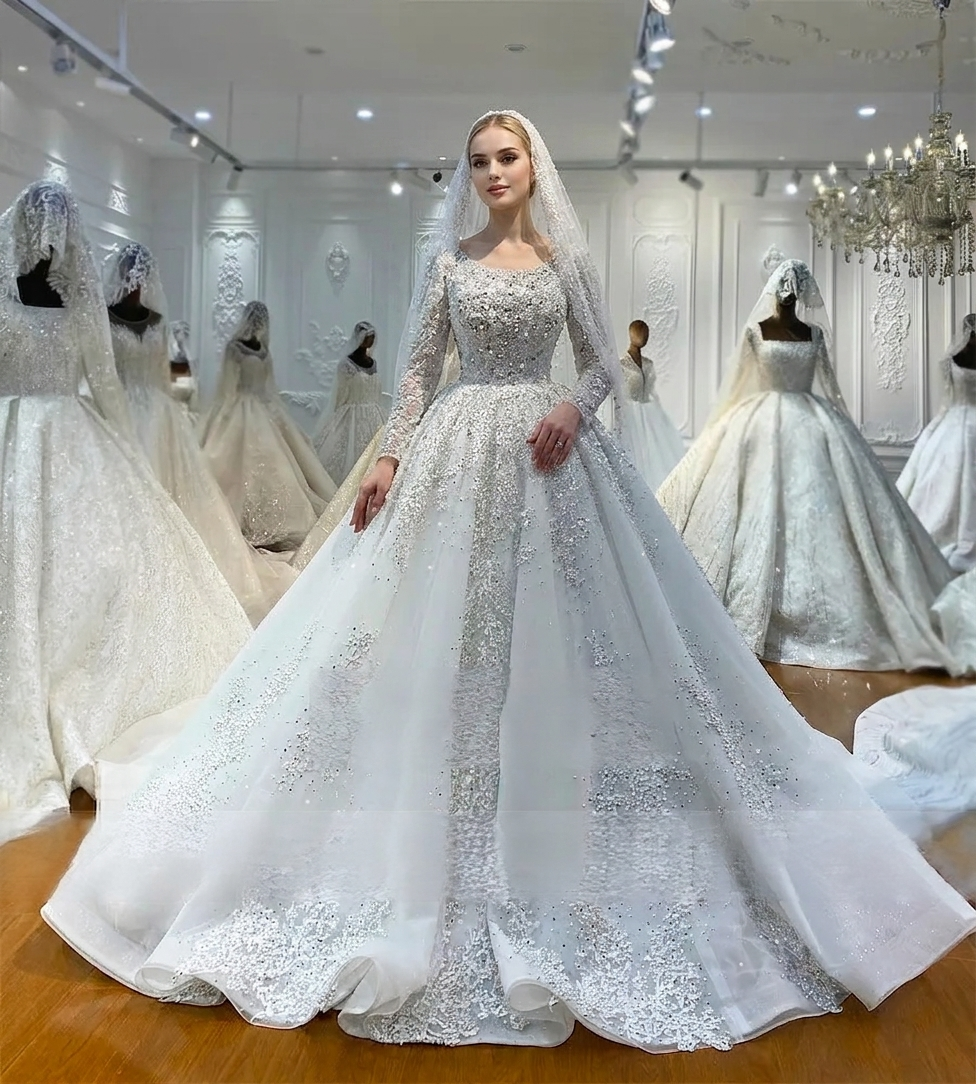 Full-length front view of an elegant model in a heavy beaded pearl wedding dress with long sleeves and a matching beaded veil, showcasing a luxury off-white ball gown silhouette in a bridal boutique.