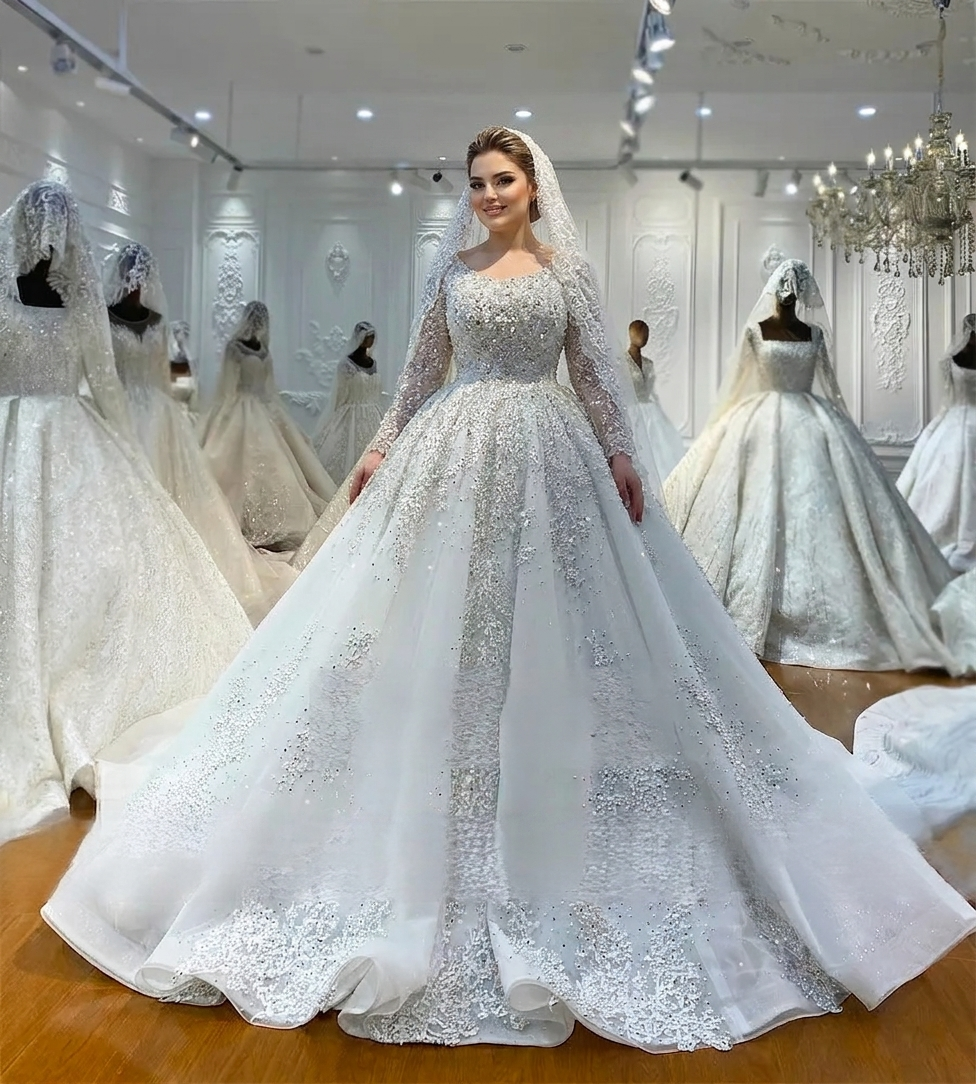 Front perspective of a model wearing a luxurious off-white ball gown featuring a round neckline, long sleeves, and intricate hand-beaded pearl and crystal embellishments throughout the bodice and skirt.