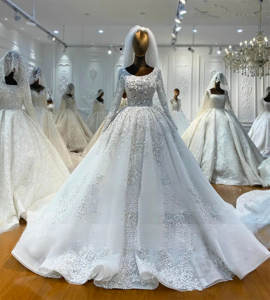 Professional showroom display of an off-white ball gown wedding dress on a mannequin, illustrating the grand volume of the skirt and the dense pearl and floral lace detailing on the long sleeves.