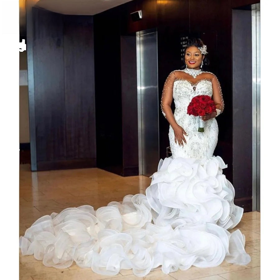 Full-length front view of an African bride in a luxury high-neck beaded mermaid wedding dress featuring sheer pearl-encrusted sleeves and a massive, dramatic ruffled organza train.