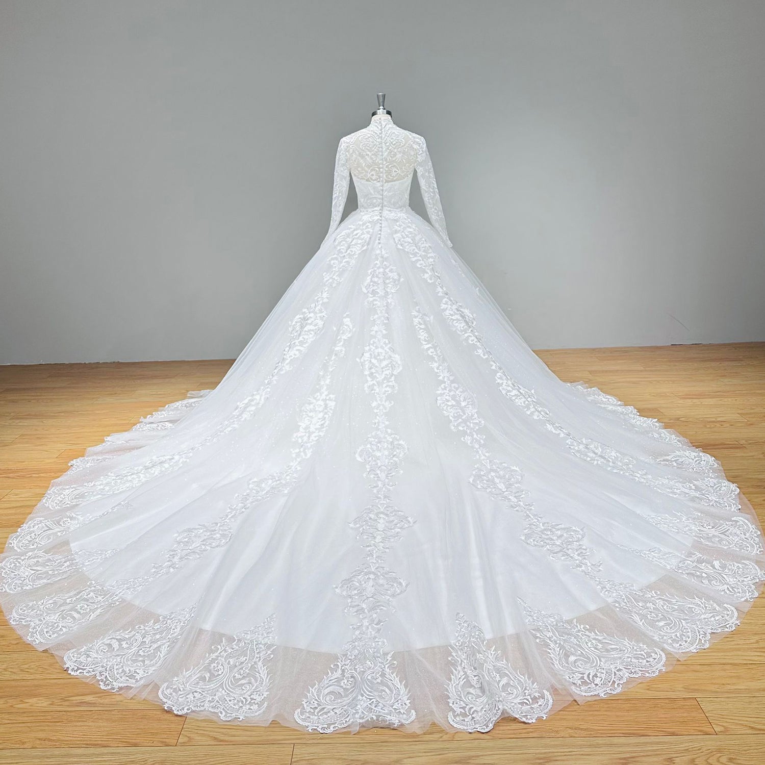 Full back view of a long-sleeve A-line wedding gown highlighting the illusion lace back, covered buttons, and a floor-length tulle pool train.