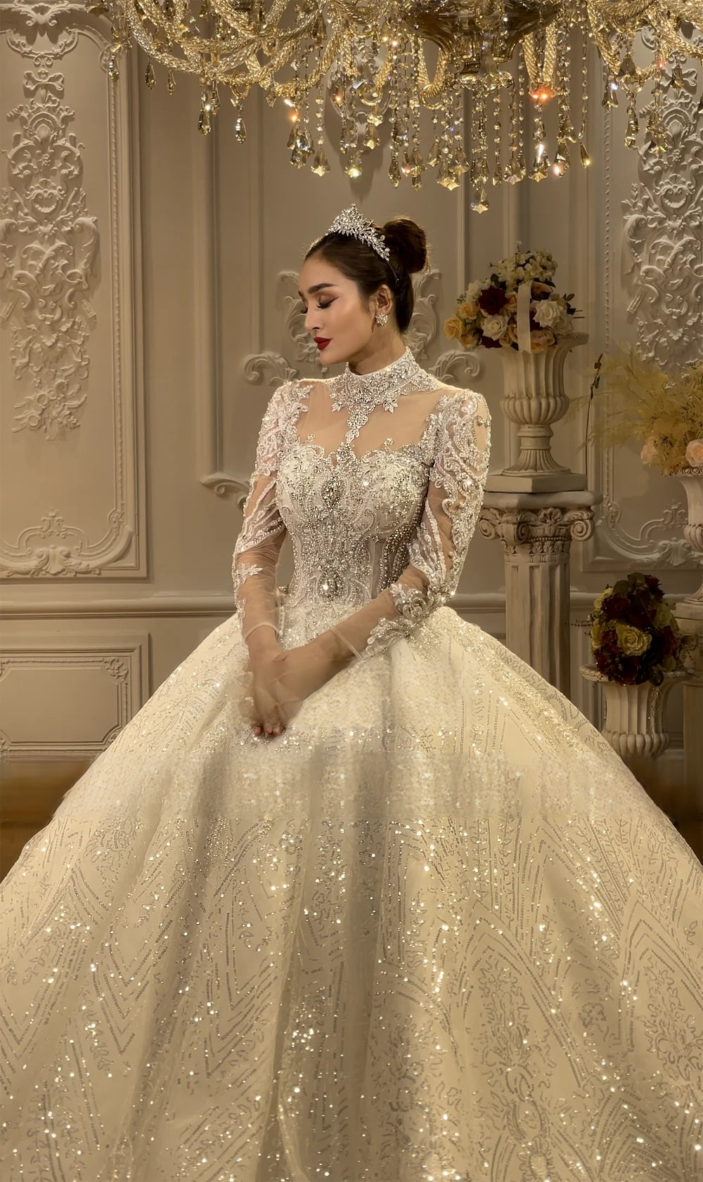 Medium-length portrait of a bride in an elegant high neck lace ball gown with long sleeves and a regal silver tiara in a luxury bridal showroom.