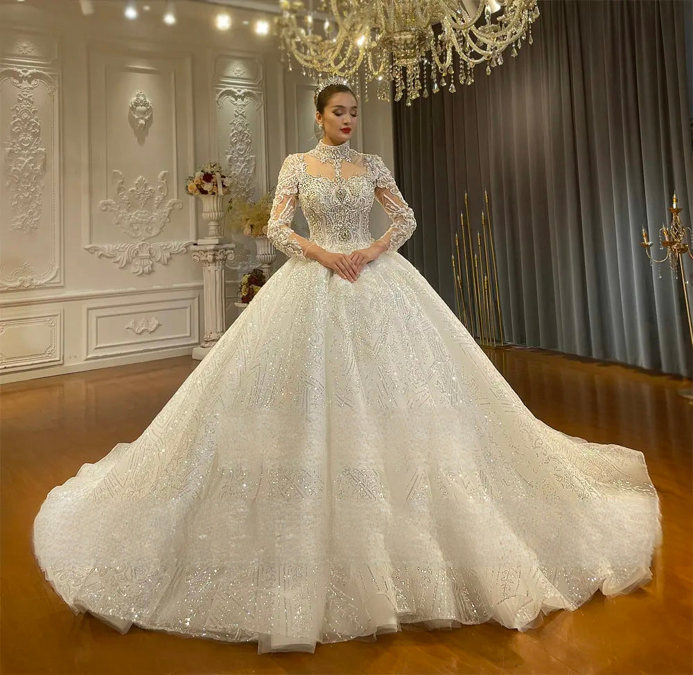 Regal perspective of an ivory high neck wedding dress with a puffy ball gown skirt and all-over silver sequin and lace appliques under a crystal chandelier.