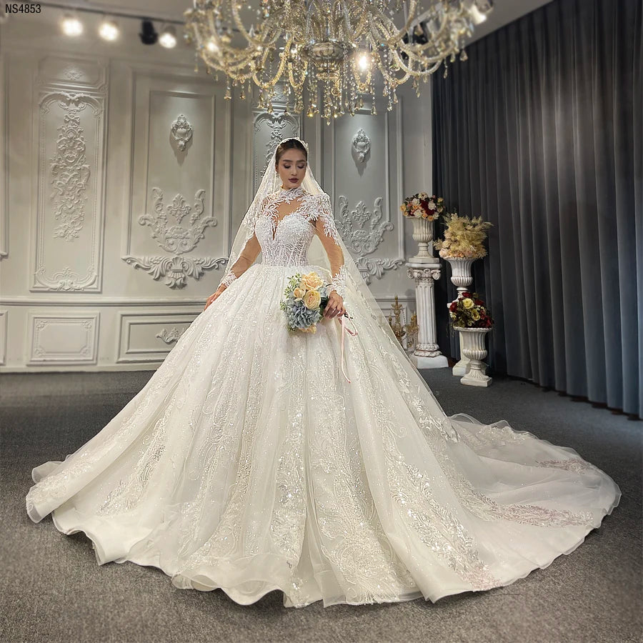 Full-length front view of an elegant bride wearing a high neckline lace wedding dress with a voluminous ball gown silhouette and long sheer sleeves featuring intricate beading.