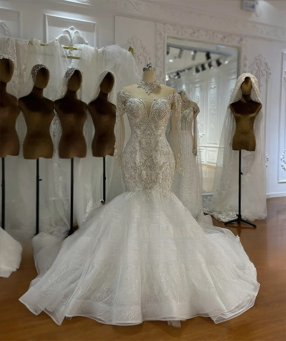 Angled front view of an off-the-shoulder long-sleeved mermaid wedding dress on a mannequin, showcasing the luxury beadwork and fitted silhouette with a lace train.
