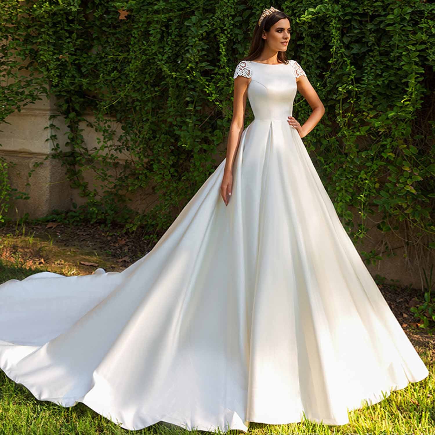 Elegant model posing in a structured white satin bridal gown with a full pleated skirt and a dramatic train against a lush green ivy wall backdrop.