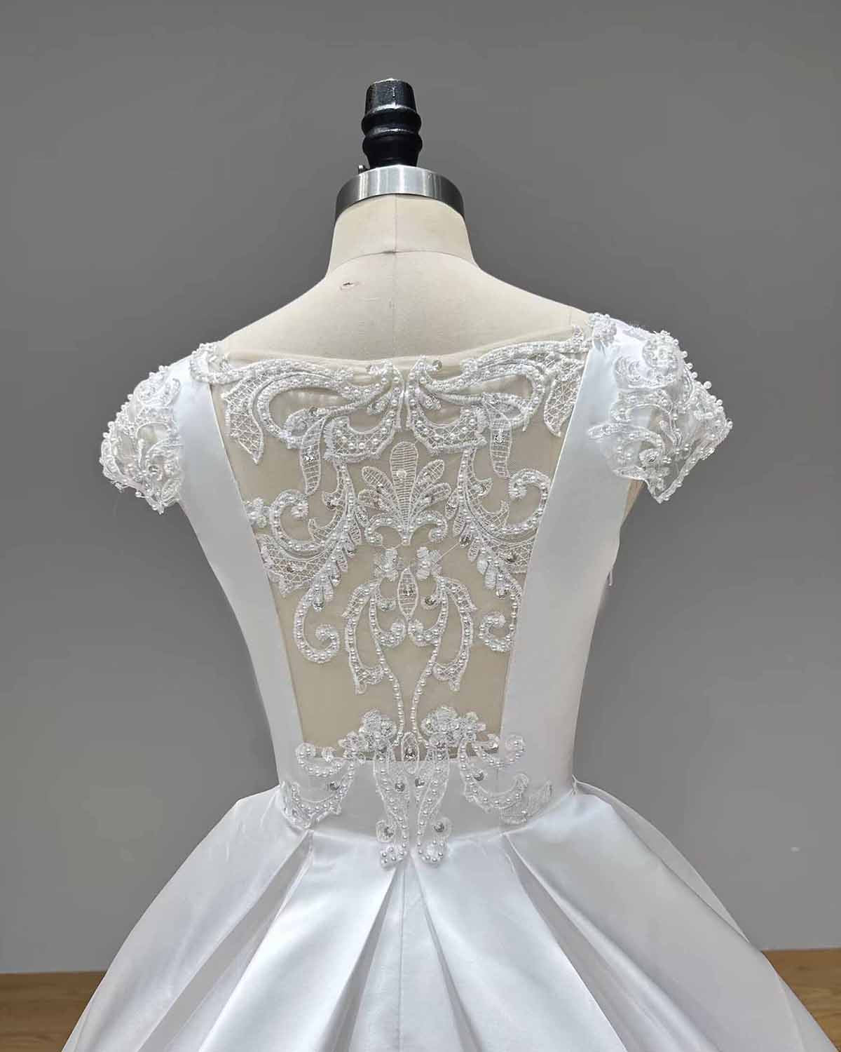 Close-up macro shot of an illusion back on a white satin bridal gown highlighting the intricate lace appliqué motifs, pearl accents, and satin-covered buttons.