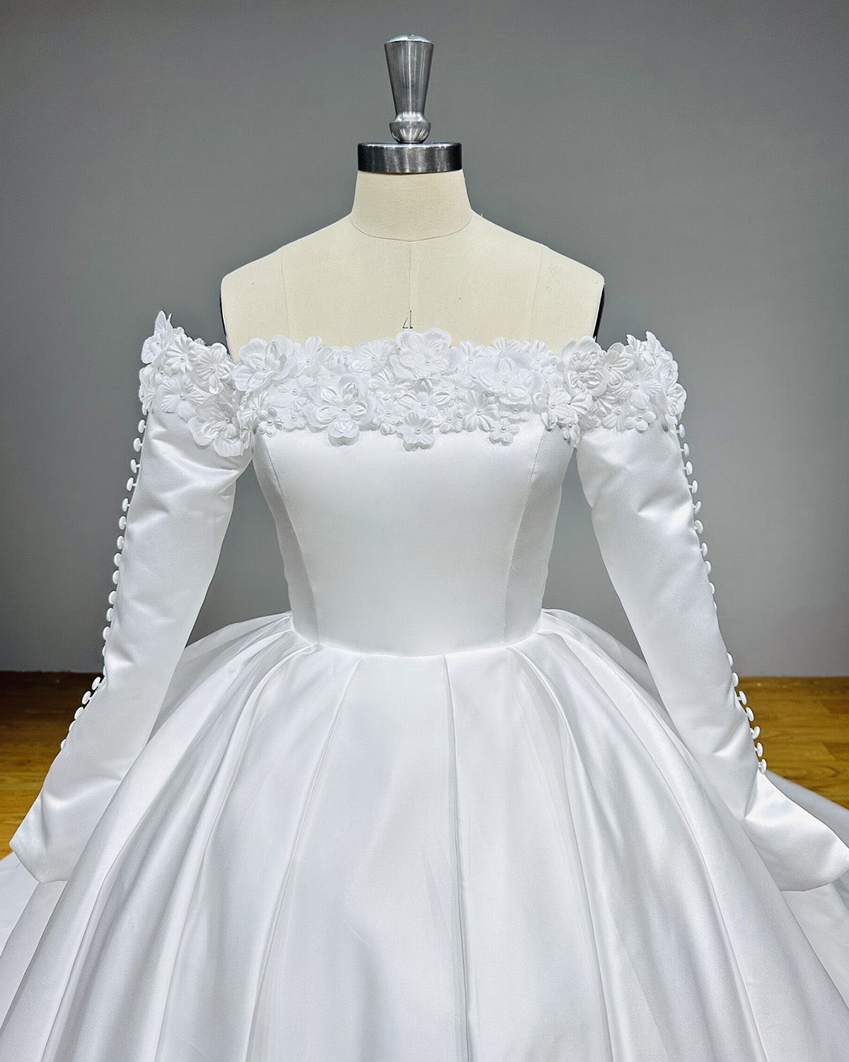 Detailed front view of a structured white satin wedding dress bodice highlighting the off-the-shoulder long sleeves decorated with fabric covered buttons.